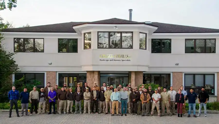 A large group of people stand together outside a two-story office building with a sign reading Hellerus, Hardware and Masonry Specialists. The group is posing for a photo on a paved area in front of the entrance.