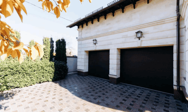 A modern house with two closed black garage doors, beige stone walls, a tiled driveway, green bushes, and yellow autumn leaves hanging from a tree in the foreground.