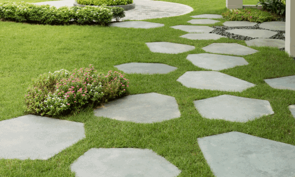 Large, irregularly shaped stone pavers form a winding pathway through a neatly manicured green lawn, bordered by small flowering bushes and trimmed shrubs, in a landscaped garden setting.