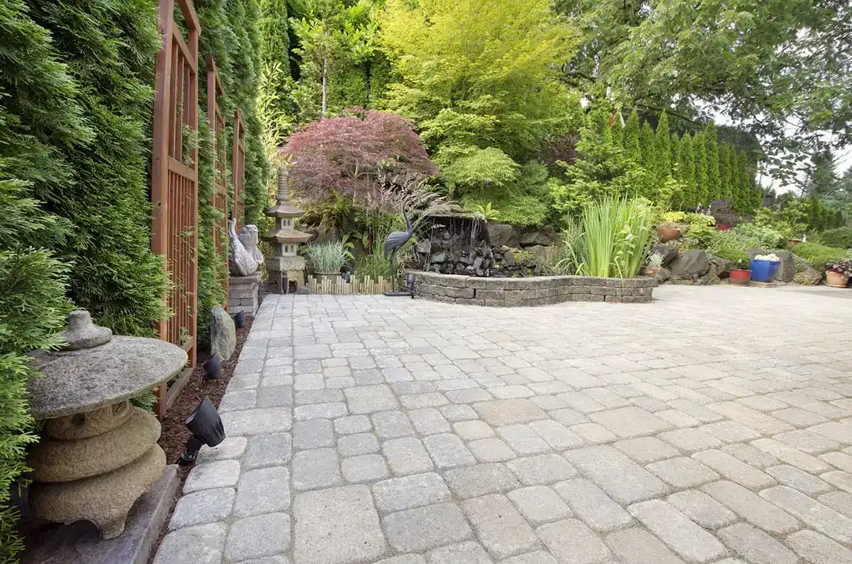 A serene backyard garden with stone pavers, lush green plants, a small waterfall, and a stone lantern. Wooden trellis and manicured trees add to the peaceful, landscaped atmosphere.