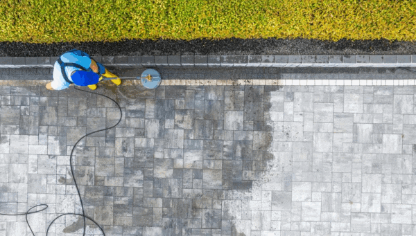 A person in blue clothing and yellow boots uses a power washer to clean a large stone-paved patio. The clean and dirty sections are clearly visible. A strip of green hedges borders the top of the patio.