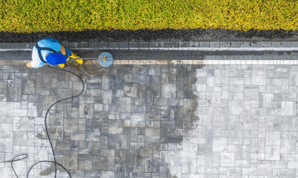 A person in blue clothing and yellow boots uses a power washer to clean a large stone-paved patio. The clean and dirty sections are clearly visible. A strip of green hedges borders the top of the patio.
